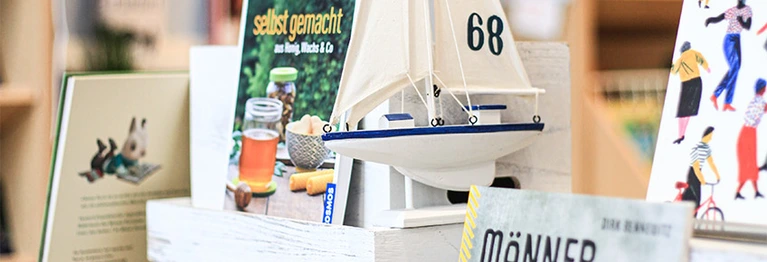 Nahansicht eines Büchertisches und einiger präsentierter Buchtitel. Ein kleines Segelschiff ist zu Dekorationszwecken auch auf dem Bild zu sehen. Die Segel sind weiß. Auf dem rechten Segel ist die Zahl 68 in dunkelbrau angebracht.