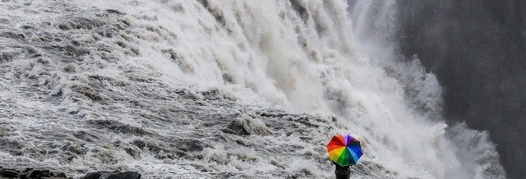 Ein Mensch steht am Rande eines Wasserfalls (schwarz-weiß) mit Regenschirm in Regenbogenfarben