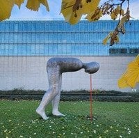 Steinskulptur Mensch beucht sich im 90 Grad l nach vorne. Steht im Freien auf einer Wiese vor dem Museumsgebäude