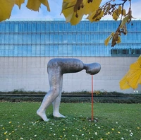 Steinskulptur Mensch beucht sich im 90 Grad l nach vorne. Steht im Freien auf einer Wiese vor dem Museumsgebäude