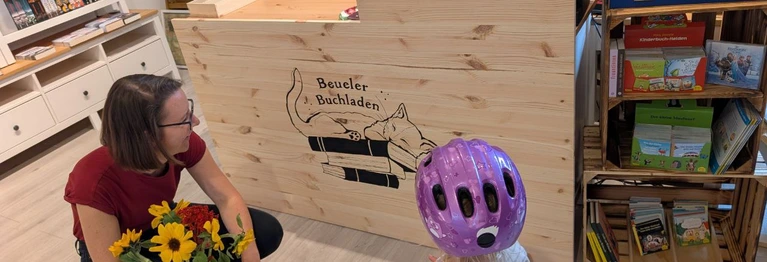 Die Inhaberin Viviane Richart mit einer jungen Kundin vor dem Logo des Beueler Buchladens am Tresen.